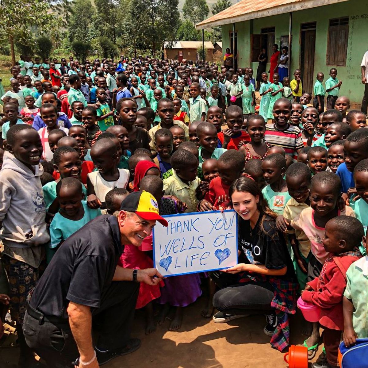 Keleigh Sperry, her dad, and many Ugandan children posing for a photo shoot while holding Thank You Wells of Life banner.